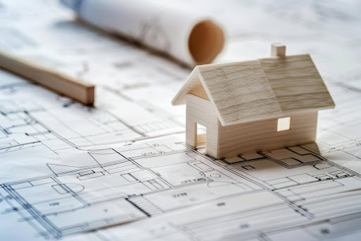 Small wooden house model sitting on architectural blueprints with a pencil nearby.